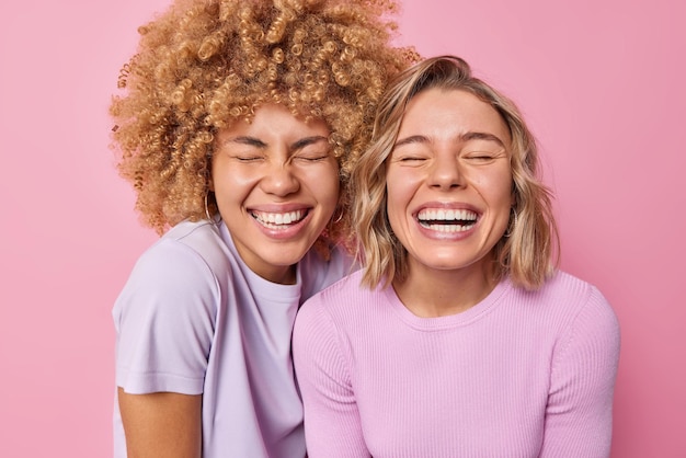 Alegres e sinceras mulheres bonitas com cabelo loiro sorriem amplamente se sentem muito felizes por manter os olhos fechados se sentem otimistas vestidas com roupas casuais isoladas sobre fundo rosa conceito de emoções humanas felizes