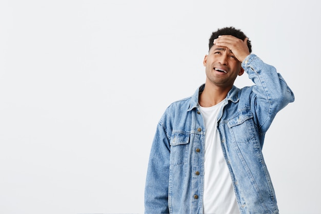 Alguém por favor faça isso em vez de mim. Retrato de homem infeliz de pele bronzeada, com cabelos cacheados escuros na camiseta branca e jaqueta azul, segurando a mão na testa, vai chorar por estar cansado do trabalho.