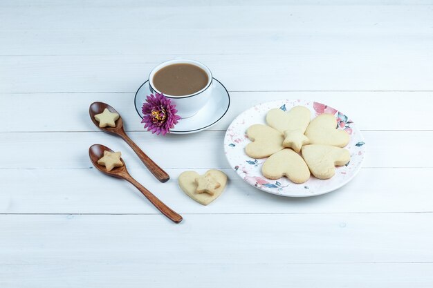 Alguns biscoitos em forma de coração e estrela com flores, biscoitos em colheres de madeira, xícara de café em um prato branco sobre fundo branco de madeira, vista de alto ângulo.