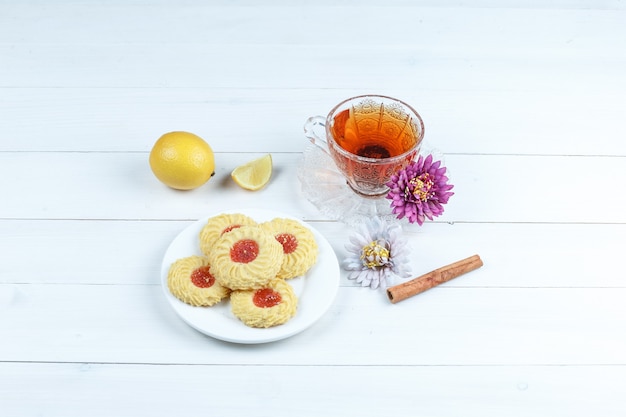 Alguns biscoitos, xícara de chá com canela, limão, flores sobre fundo branco de placa de madeira, vista de alto ângulo.