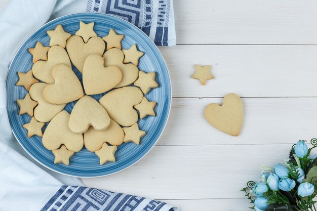 Alguns cookies com flores em um prato sobre fundo de madeira e toalha de cozinha, plana leigos.
