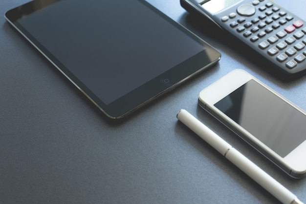 Alguns dispositivos eletrônicos indicados no fundo cinzento. Smart phone, pad e calculadora, todos digitais, exceto uma caneta. Local de trabalho da cena.