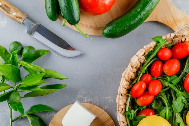 Alguns tomates com faca, limão, pepino, queijo, verduras em uma tábua na superfície cinza, vista superior