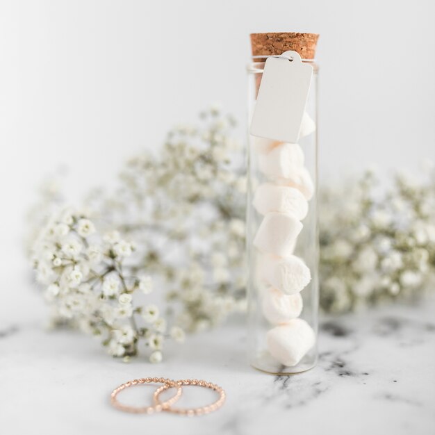 Alianças de casamento; tubos de ensaio de marshmallow com tag e flores da respiração do bebê no plano de fundo texturizado