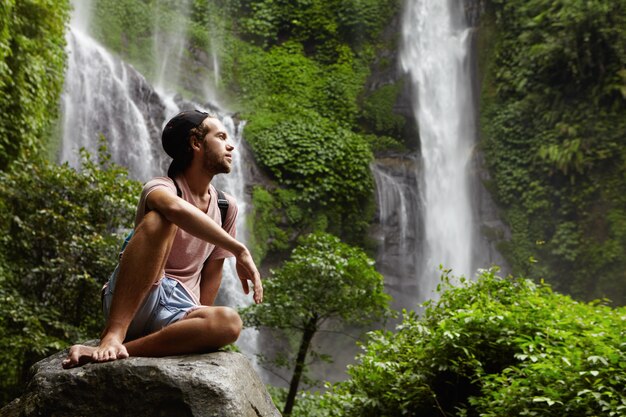 Alpinista caucasiano elegante vestido casualmente sentado com os pés descalços em uma pedra grande e relaxando durante uma longa e difícil jornada na floresta tropical. Homem barbudo em snapback contemplando a bela natureza ao seu redor