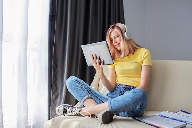 Aluna adolescente em fones de ouvido olhando na tela do tablet digital, sentado em casa no sofá. bate-papo por vídeo, aula online, e-learning, mulheres conversando e aprendendo remotamente