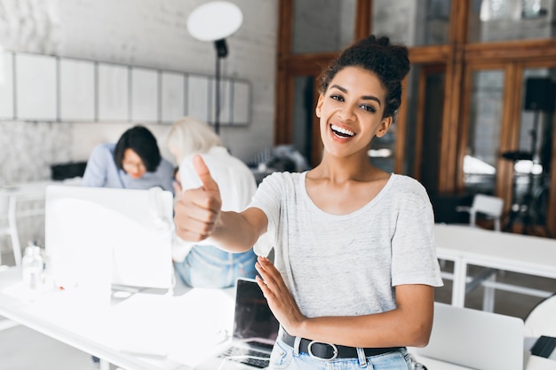 Foto grátis aluna africana alegre com penteado curto, segurando o polegar depois de passar nos exames. retrato de mulher negra feliz em t-shirt cinza se divertindo no escritório enquanto seus colegas trabalhando no projeto.