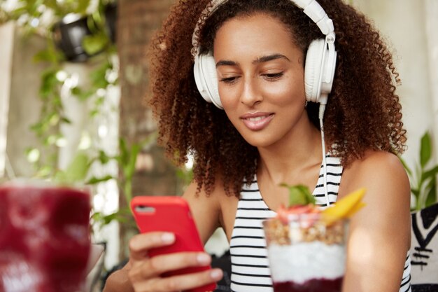 Aluna afro-americana ouve aula de áudio em fones de ouvido modernos no smartphone, conectado à internet sem fio em um café aconchegante, melhora o conhecimento da língua estrangeira. Tecnologia e juventude