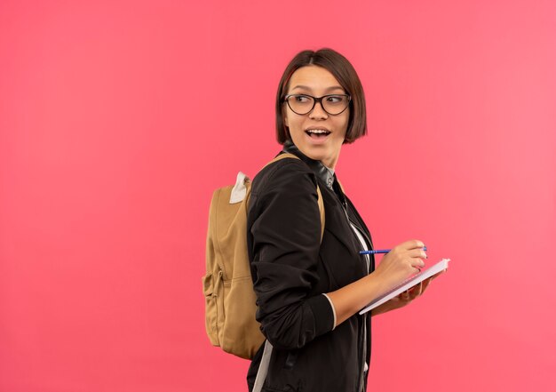 Aluna alegre de óculos e bolsa traseira em pé na vista de perfil, segurando uma caneta e um bloco de notas, olhando para trás, isolada em rosa