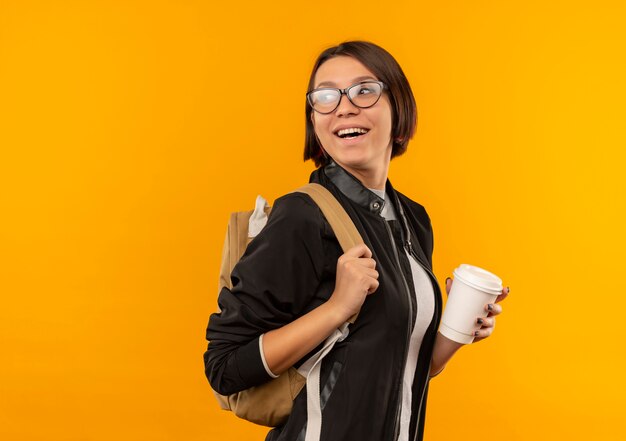 Aluna alegre de óculos e bolsa traseira em vista de perfil, olhando para trás, segurando uma xícara de café isolada em laranja
