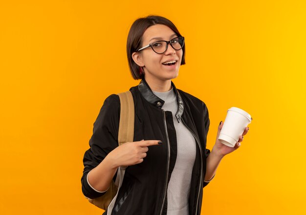 Aluna alegre de óculos e mochila segurando e apontando para uma xícara de café de plástico isolada em laranja