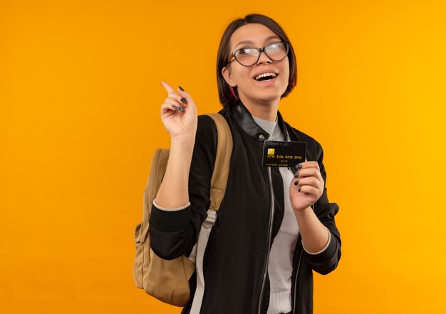 Aluna alegre de óculos e mochila segurando o cartão de crédito, olhando para o lado, levantando o dedo isolado na laranja