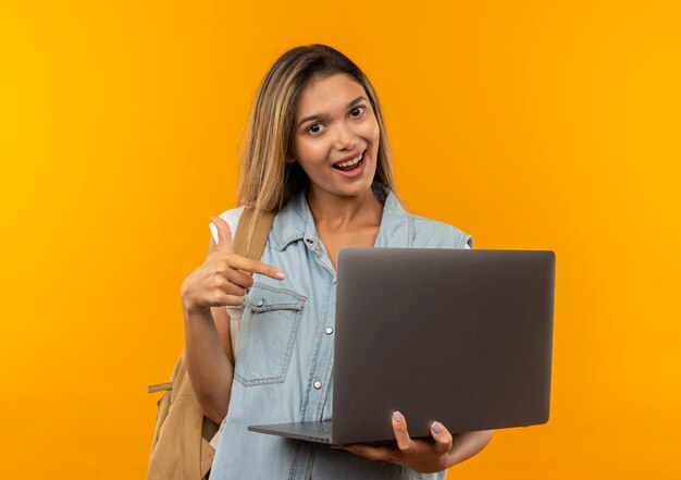 Aluna alegre e bonita vestindo uma bolsa de costas, segurando e apontando para um laptop isolado em laranja