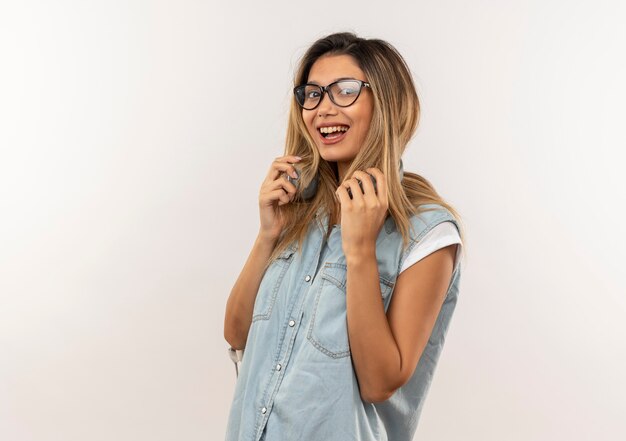 Foto grátis aluna alegre, jovem e bonita usando óculos e bolsa traseira com fones de ouvido no pescoço segurando fones de ouvido isolados no branco