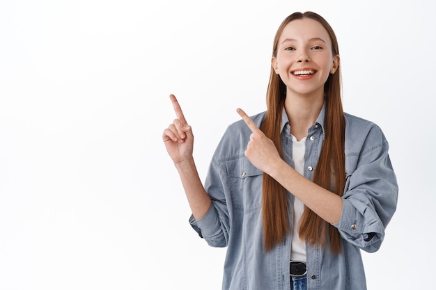 Aluna alegre, menina adolescente rindo e sorrindo, apontando o dedo de lado, mostrando o logotipo, banner com descontos especiais, oferta da loja, em pé sobre uma parede branca