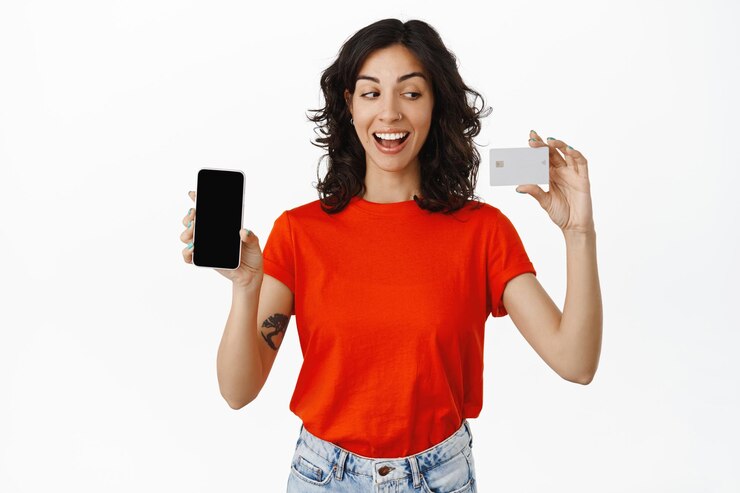 Foto grátis aluna alegre pagando on-line, mostra o cartão de crédito do banco, tela do telefone móvel, sorrindo feliz e satisfeito, comprando no aplicativo, fundo branco.