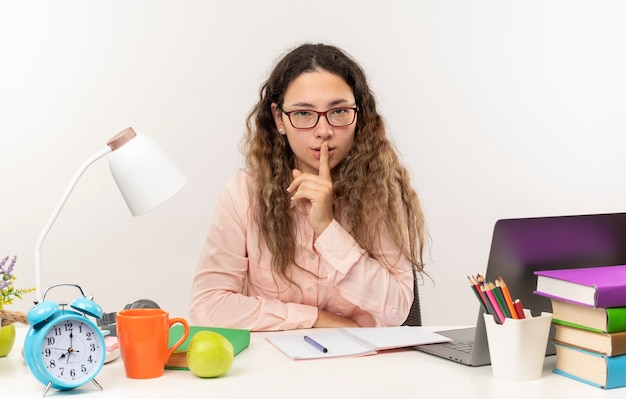 Aluna bonita e confiante usando óculos, sentada na mesa com as ferramentas da escola, fazendo sua lição de casa, gesticulando para o silêncio isolado no branco