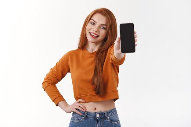 Aluna bonita e sorridente ruiva caucasiana com suéter laranja, rastreando seu pedido online, mostrando a foto legal que ela quer postar nas redes sociais, segurando um smartphone, parede branca