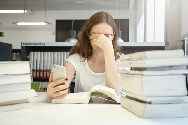 Aluna cansada da escola de economia, sentada na biblioteca, esfregando os olhos, entediada com a leitura do manual de auditoria, verificando o tempo usando seu aplicativo de telefone inteligente, cercada por pilhas de livros