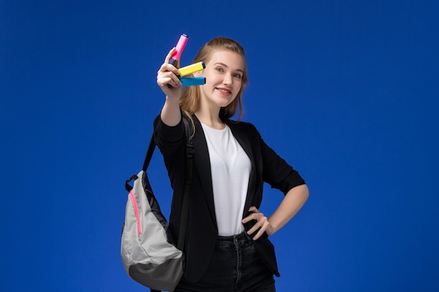 Aluna com jaqueta preta e mochila segurando canetas de feltro sorrindo na parede azul. Aluna de jaqueta preta.