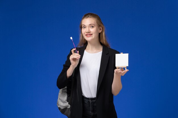 Aluna com jaqueta preta e mochila segurando o cavalete na parede azul, desenho da faculdade de arte