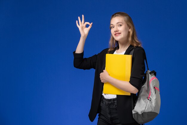 Aluna com uma jaqueta preta usando uma mochila e segurando uma pasta amarela na aula de universidade de parede azul