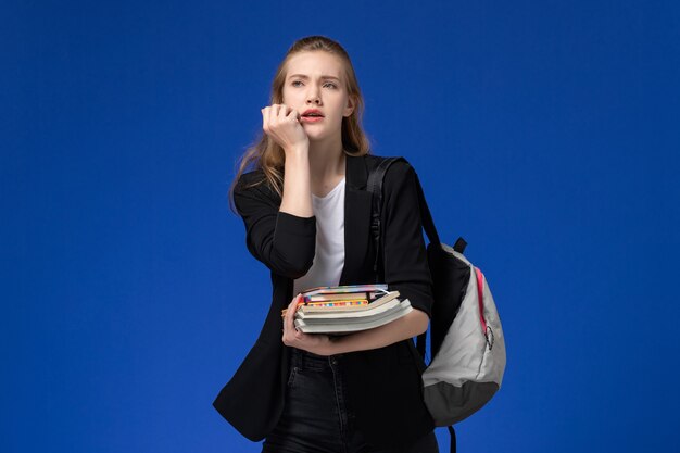 Aluna com uma jaqueta preta usando uma mochila segurando livros na parede azul claro