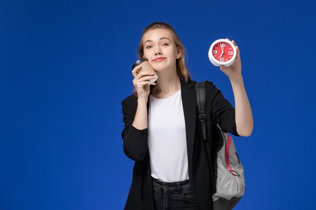 Aluna com uma jaqueta preta usando uma mochila segurando relógios e café na parede azul da universidade universitária