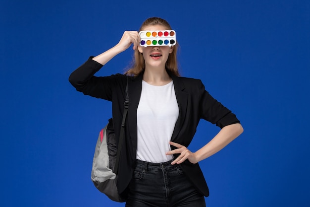 Foto grátis aluna com uma jaqueta preta usando uma mochila segurando tintas para desenhar na parede azul-clara desenho na parede azul claro.