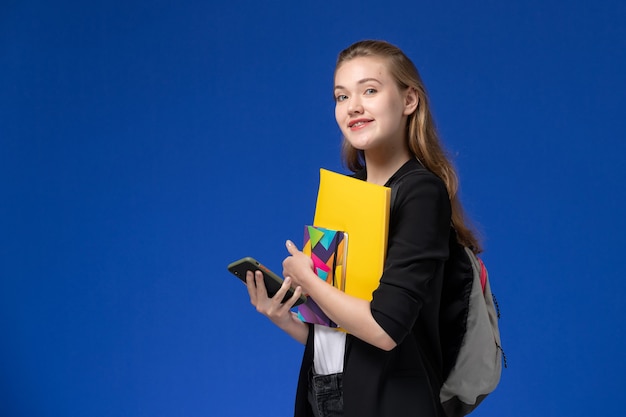 Aluna com uma jaqueta preta usando uma mochila segurando um arquivo e um caderno no livro de parede azul.