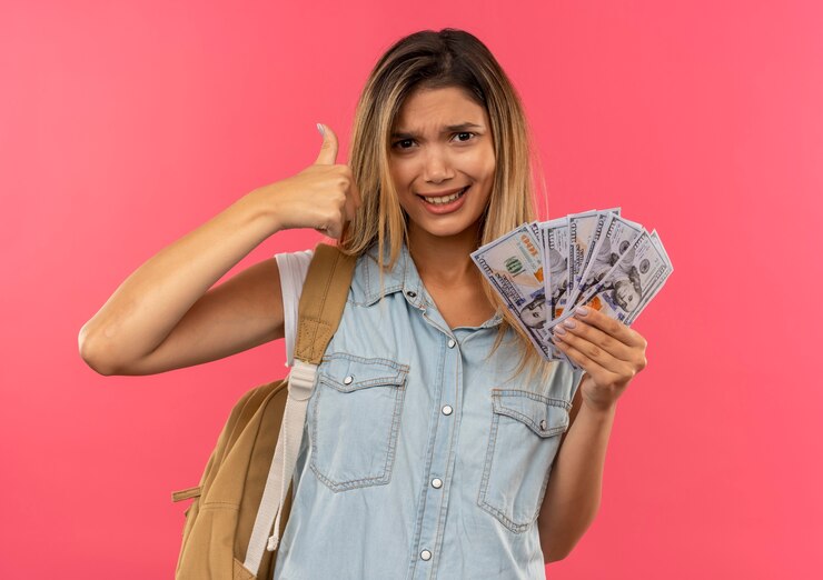 Foto grátis aluna confiante jovem e bonita usando uma bolsa de costas segurando dinheiro e aparecendo o polegar isolado na rosa