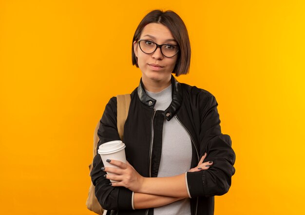 Aluna confiante usando óculos e bolsa traseira em pé com a postura fechada e segurando a xícara de café isolada em laranja com espaço de cópia