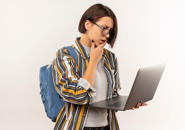 Aluna confusa usando óculos e bolsa traseira segurando e olhando para o laptop com a mão no queixo isolado no branco