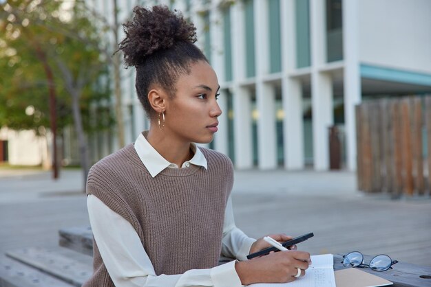 Aluna de cabelos encaracolados pensativa faz lição de casa ao ar livre anota notas no diário mantém telefone celular navega em sites usa camisa branca e colete focado na distância Estudando conceito