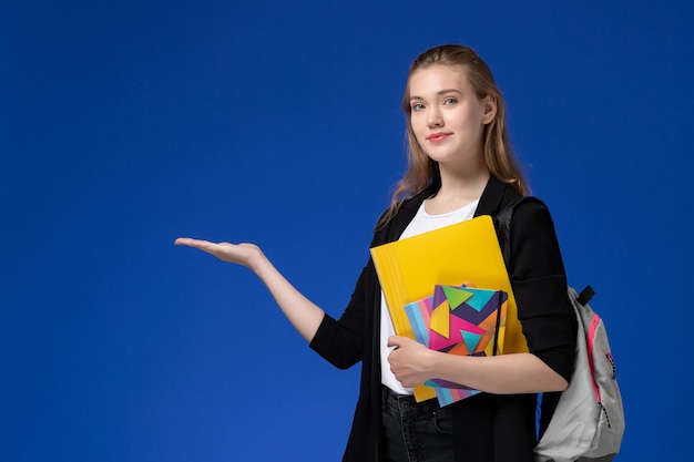 Aluna de frente para uma aluna de camisa branca e jaqueta preta, usando uma mochila, segurando arquivos com cadernos na parede azul.