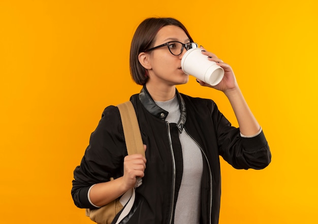 Aluna de óculos e bolsa traseira bebendo café de uma xícara de café de plástico olhando para o lado isolado em fundo laranja