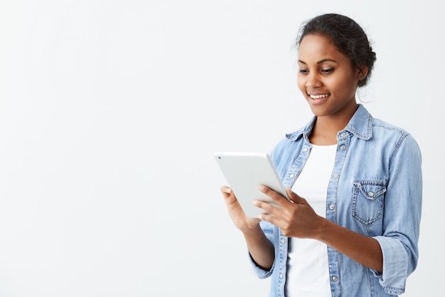 Aluna de pele escura jovem alegre com sorriso bonito em pé na parede branca, usando o tablet, verificando o feed de notícias em suas contas de rede social. garota muito afro-americana, navegar na internet em t