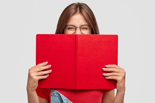 Aluna feliz ri positivamente, usa óculos redondos, se esconde atrás de um livro vermelho, sorri ao ler algo engraçado, posa contra uma parede branca. Conceito de pessoas, juventude, educação e leitura
