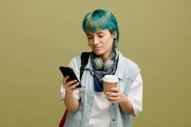 Aluna insegura usando fones de ouvido e bandana no pescoço e mochila segurando a xícara de café de papel usando telefone celular isolado em fundo verde-oliva