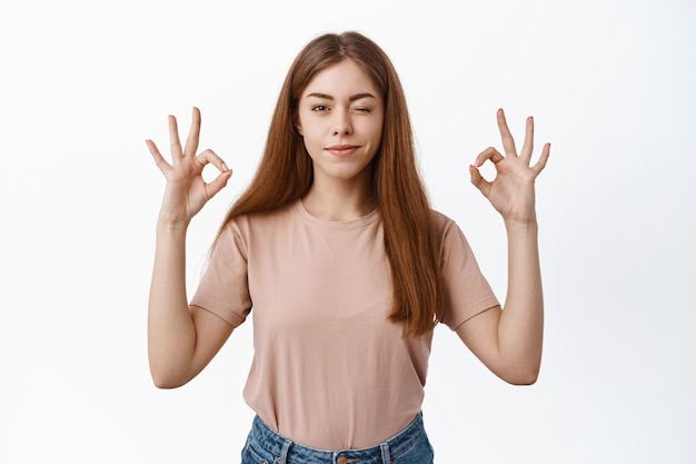 Aluna jovem confiante piscando, sorrindo e dando sinais de ok, diga sim, recomende um bom produto, aprove ou elogie sua escolha, parada determinada sobre uma parede branca