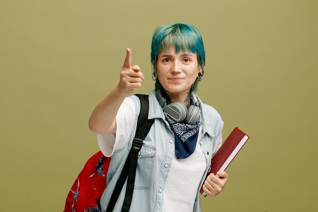 Aluna jovem descontente usando fones de ouvido e bandana no pescoço e mochila segurando o caderno olhando para a câmera levantando o dedo isolado no fundo verde oliva