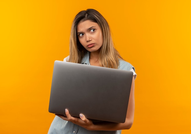 Aluna jovem e bonita confusa usando uma bolsa de costas segurando um laptop e olhando para o lado isolado em laranja com espaço de cópia