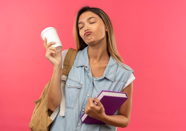 Foto grátis aluna jovem e bonita satisfeita com uma bolsa nas costas segurando um livro e uma xícara de café de plástico com os olhos fechados, isolada na parede rosa