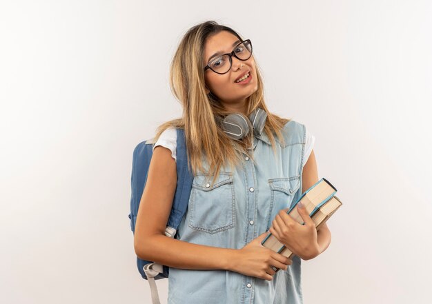 Aluna jovem e bonita satisfeita usando óculos e bolsa traseira com fones de ouvido no pescoço segurando livros isolados na parede branca