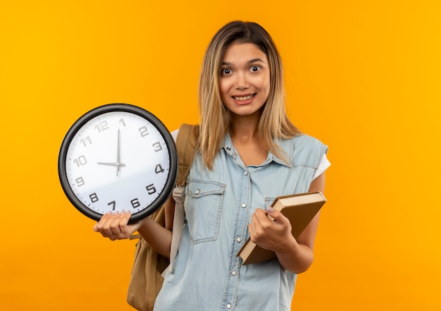 Foto grátis aluna jovem e bonita sorridente usando uma bolsa de costas segurando um livro e um relógio isolado na parede laranja