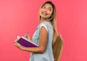 Foto grátis aluna jovem e bonita sorridente usando uma bolsa traseira em pé na vista de perfil, segurando um livro aberto e uma caneta, olhando para trás, isolada na parede rosa
