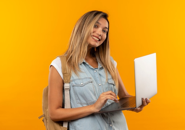 Foto grátis aluna jovem e bonita sorridente vestindo uma bolsa de costas segurando e usando um laptop isolado na parede laranja