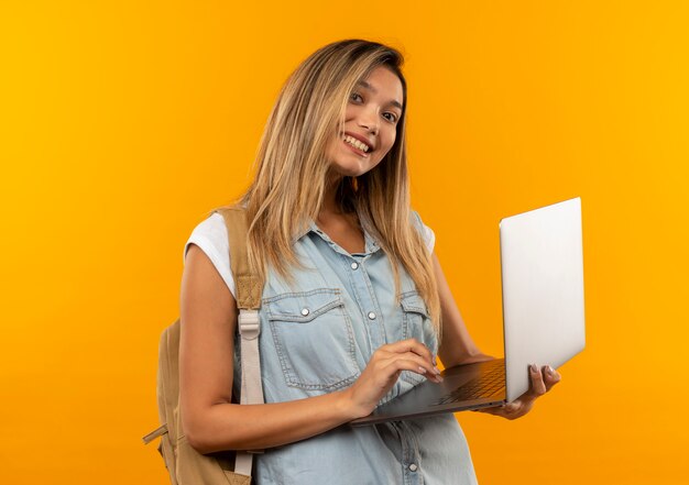 Aluna jovem e bonita sorridente vestindo uma bolsa de costas segurando e usando um laptop isolado na parede laranja