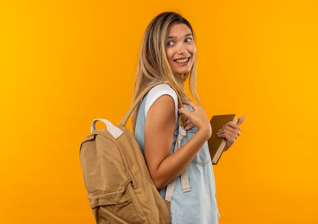 Aluna jovem e bonita sorridente, vestindo uma bolsa traseira em pé na vista de perfil, segurando o livro e olhando para trás, isolada na parede laranja