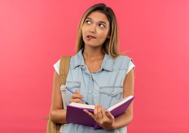 Aluna jovem e pensativa usando uma bolsa de costas segurando um livro aberto e uma caneta olhando para o lado mordendo o lábio isolado na parede rosa